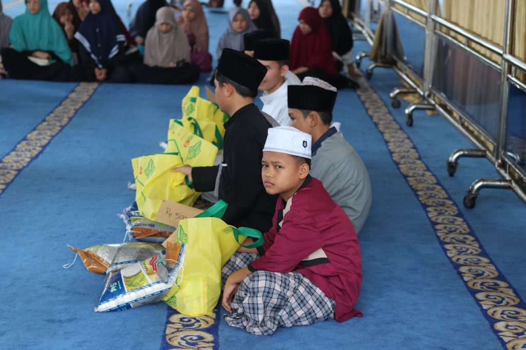 people-sitting-on-a-carpet-with-sack-in-front-of-them-12028608 Boys attending a religious gathering in Bengkulu, Indonesia, sitting with food packages.
